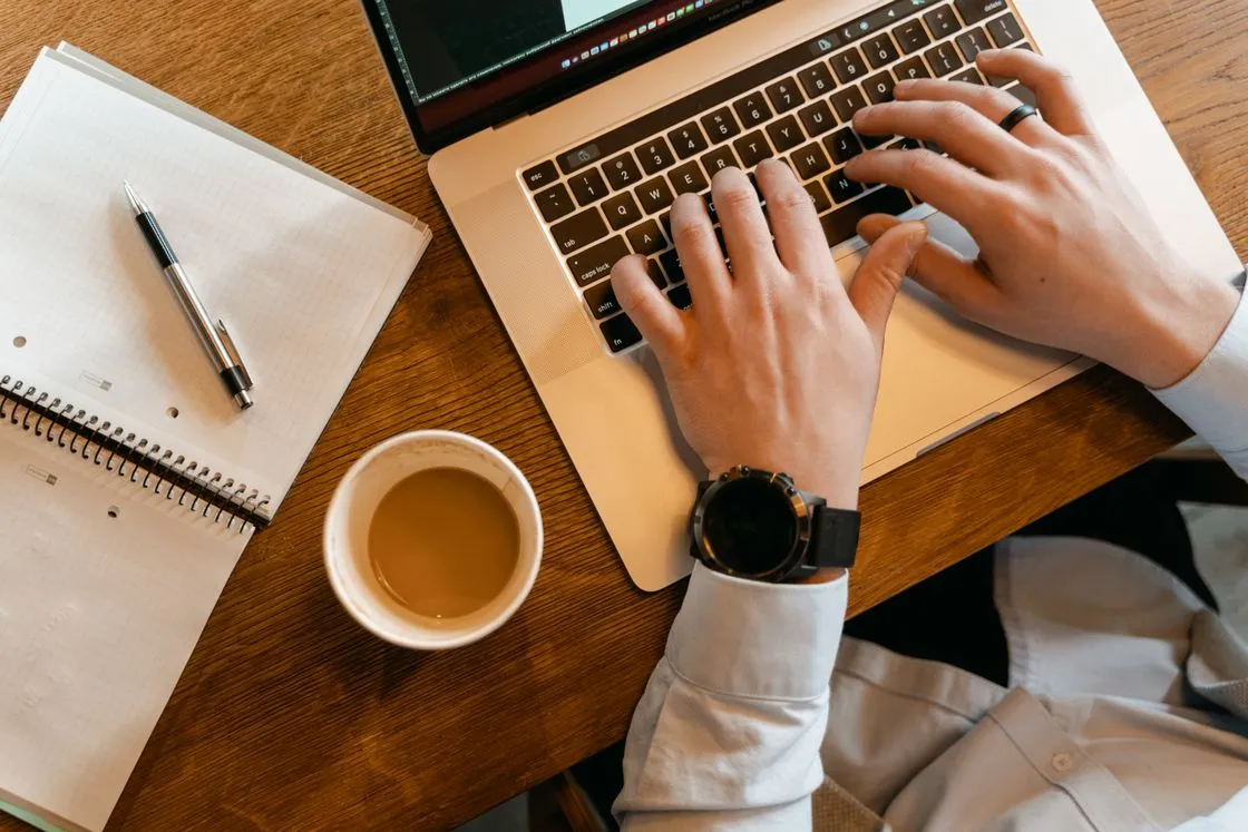 Hands typing on a laptop beside a cup of coffee and an open notebook on a wooden desk