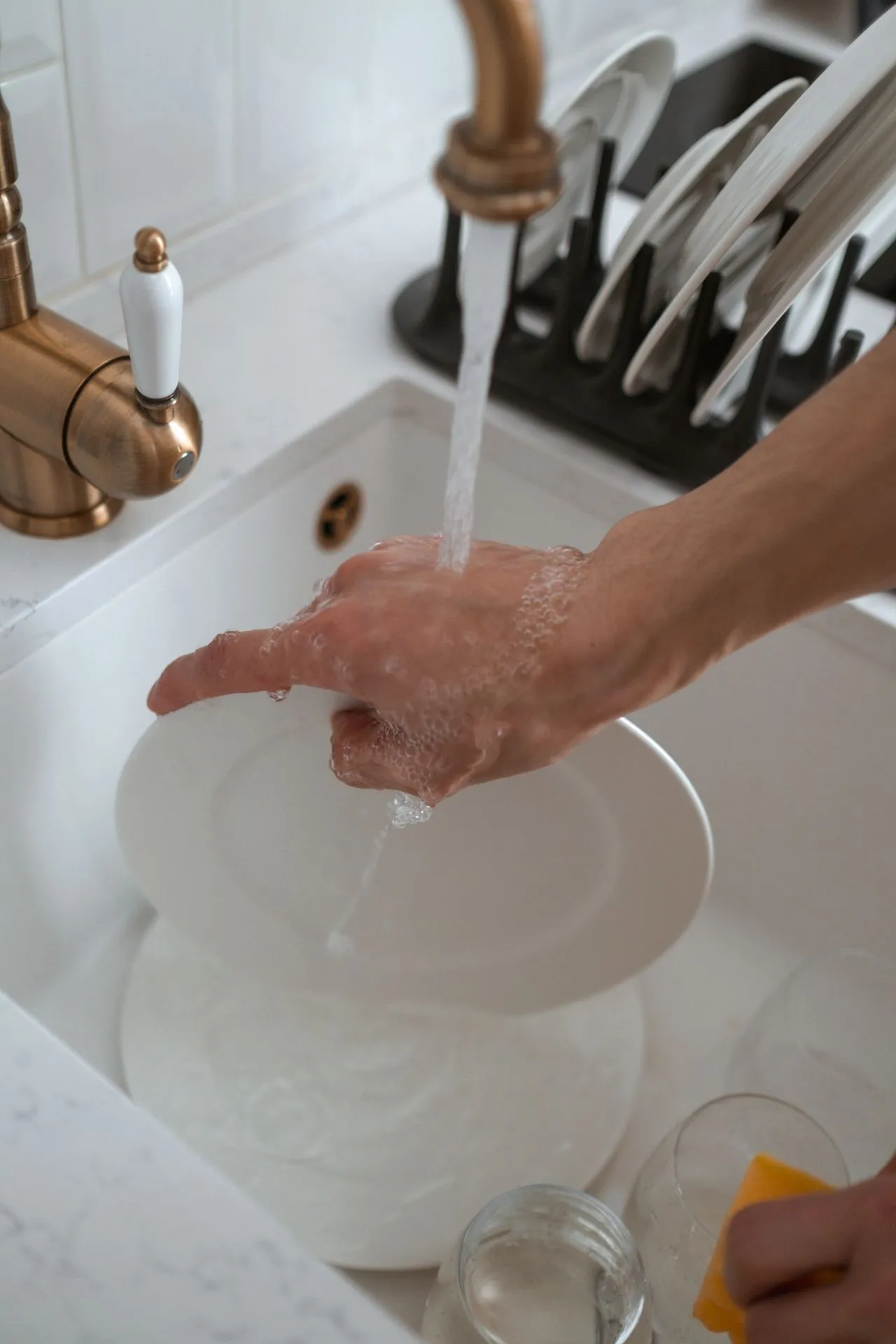 Hand rinsing a white plate under a running tap at a kitchen sink